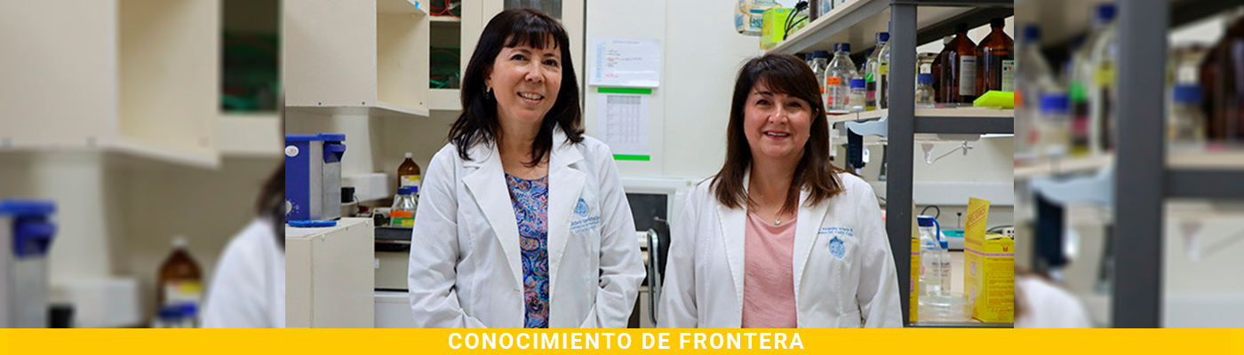 Dos mujeres científicas con batas blancas sonríen en un laboratorio, frente a estantes con equipos